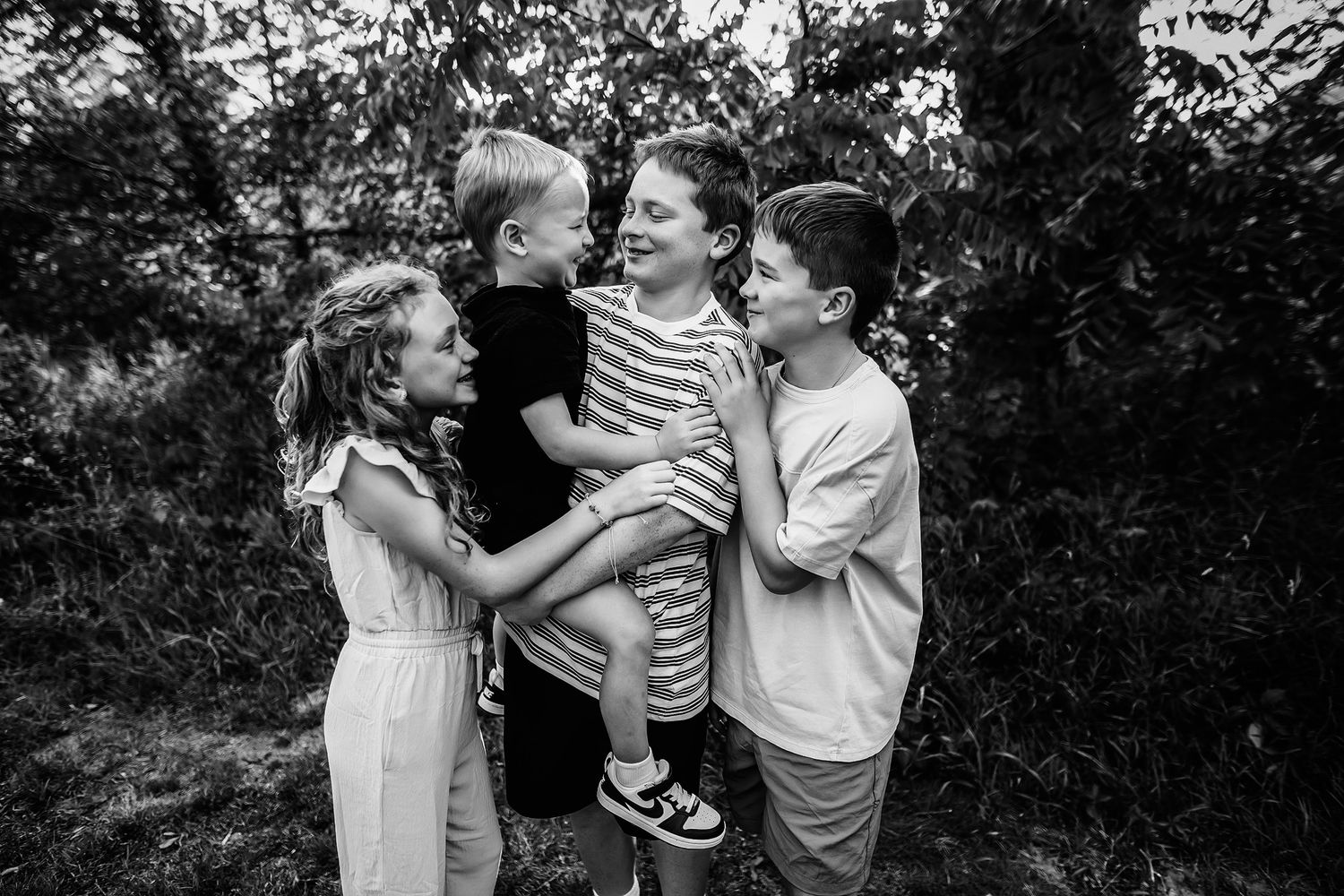 Black and white photo of four happy children hugging and laughing together outdoors among trees.