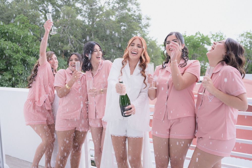 Bride in white outfit pops champagne with bridesmaids in matching pink pajamas celebrating outdoors.