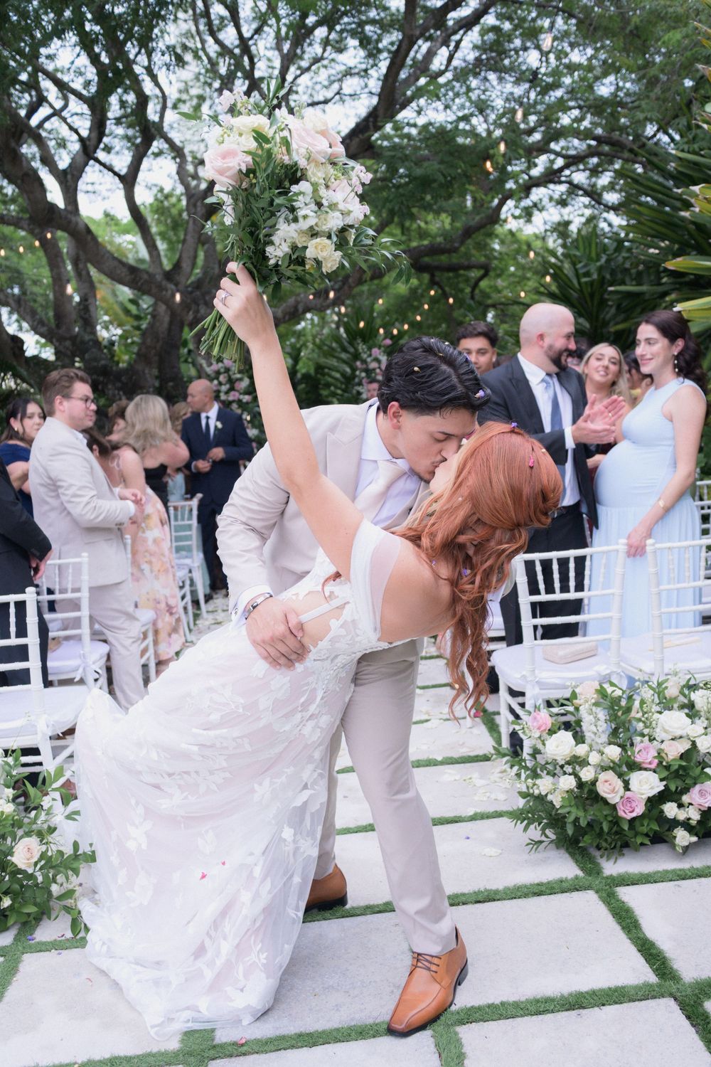Groom dips bride for a kiss at outdoor wedding ceremony while guests applaud, bride holding bouquet aloft.