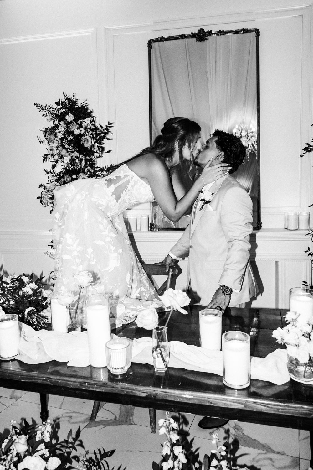Black and white photo of bride and groom sharing a kiss at decorated wedding altar with candles and floral arrangements.