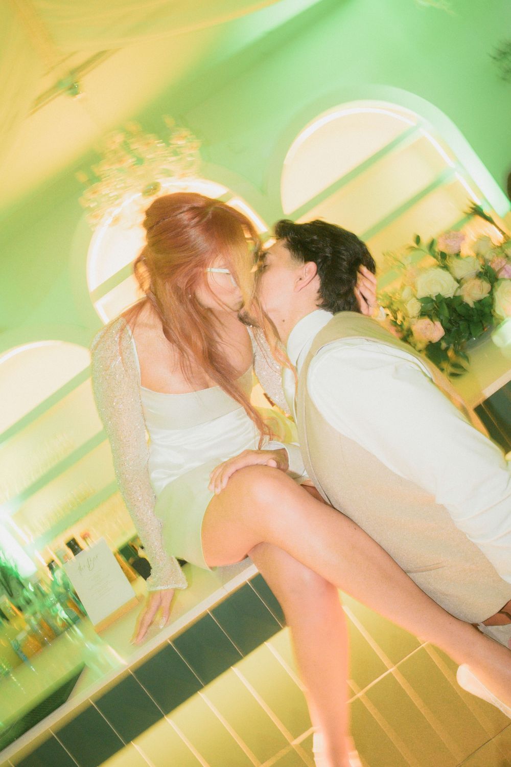 A couple shares a romantic kiss at a wedding reception in a green-lit venue with floral arrangements.