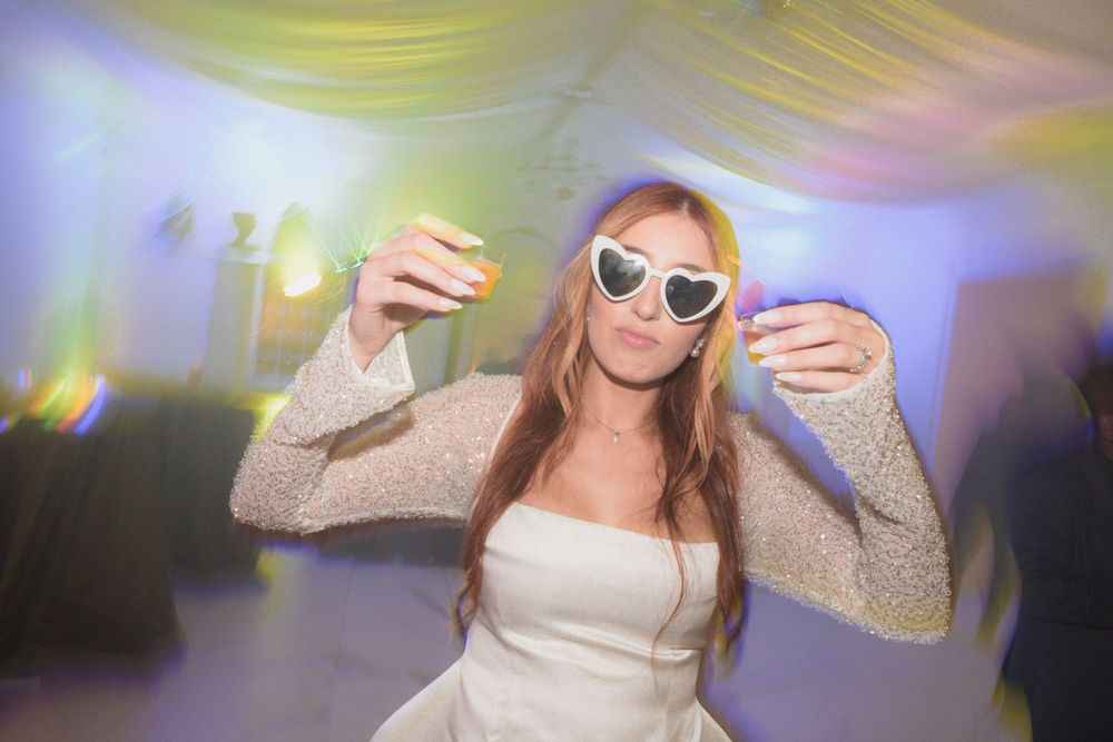 Bride dancing at wedding reception wearing white dress and heart-shaped sunglasses under colorful lights.