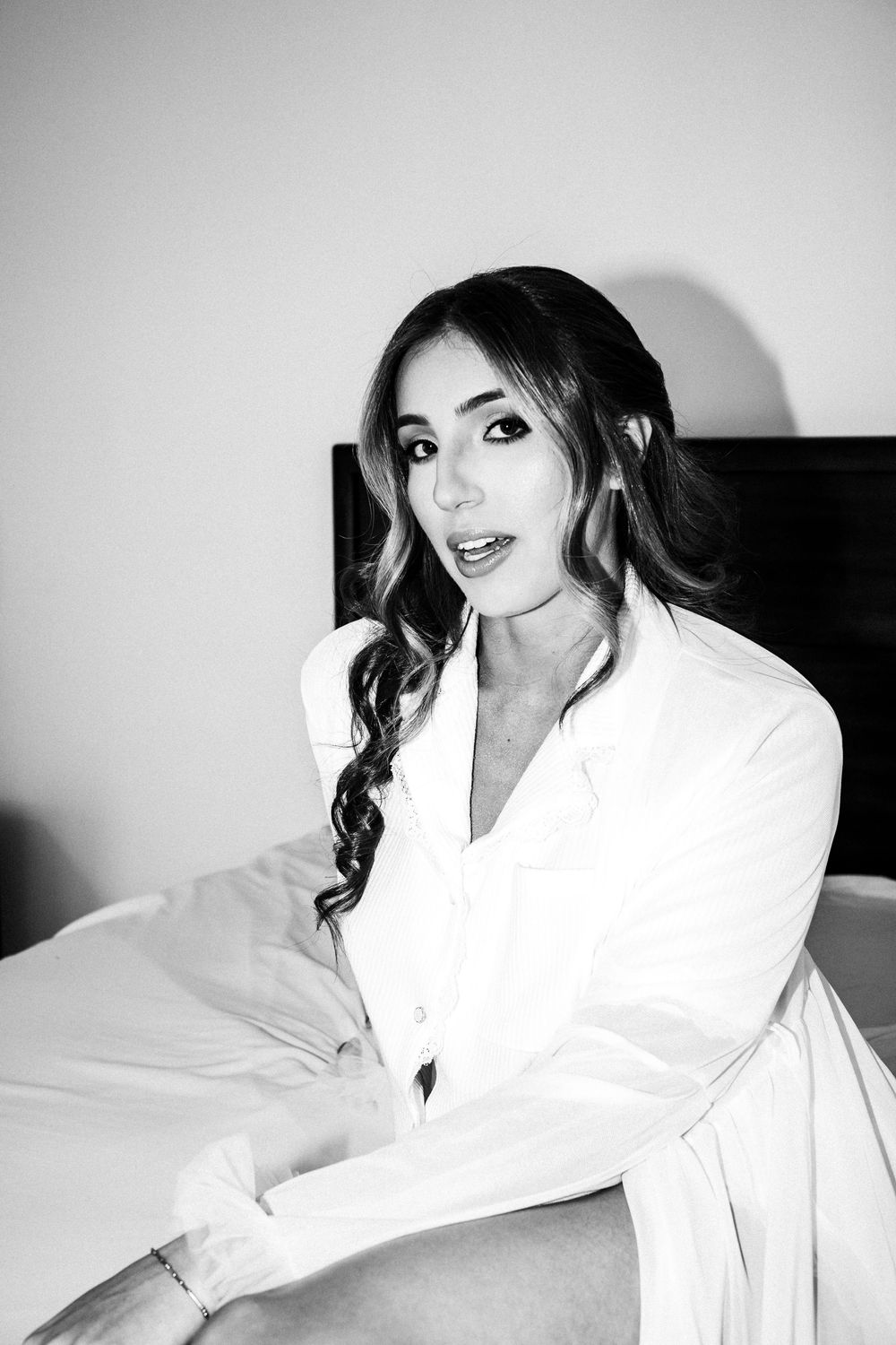 Black and white portrait of a young woman with long wavy hair sitting on a bed wearing a white blazer.