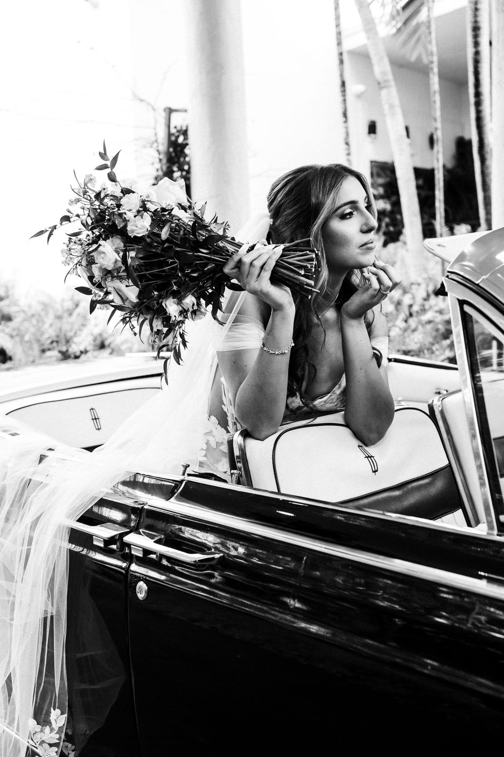 Elegant bride holding a floral bouquet while leaning out of a vintage classic car, black and white wedding photography.