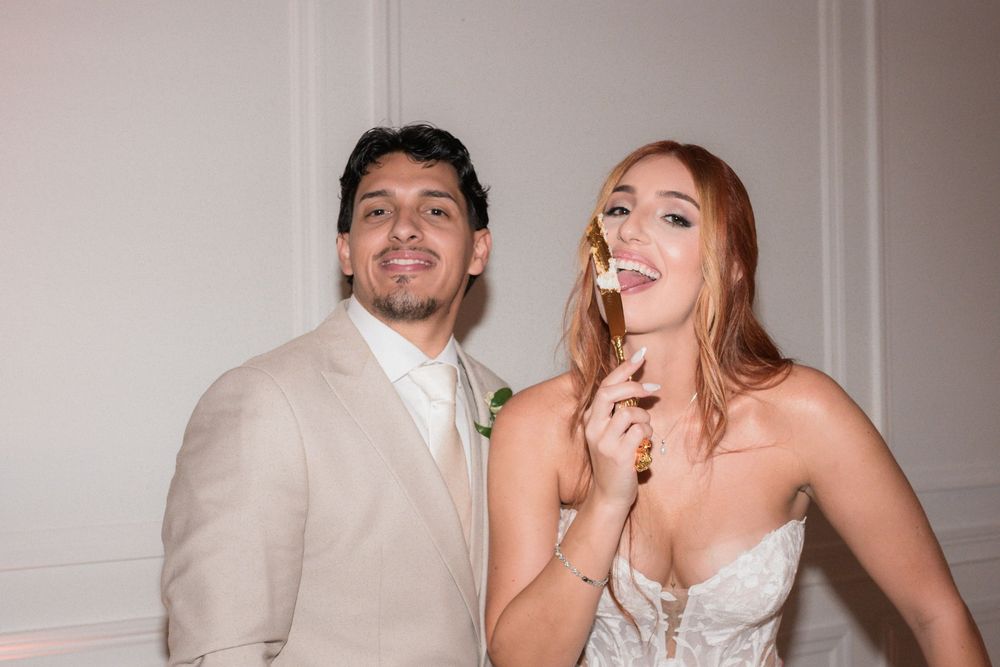 Bride and groom posing playfully at wedding reception, woman in lace gown holding cake, man in beige suit smiling.