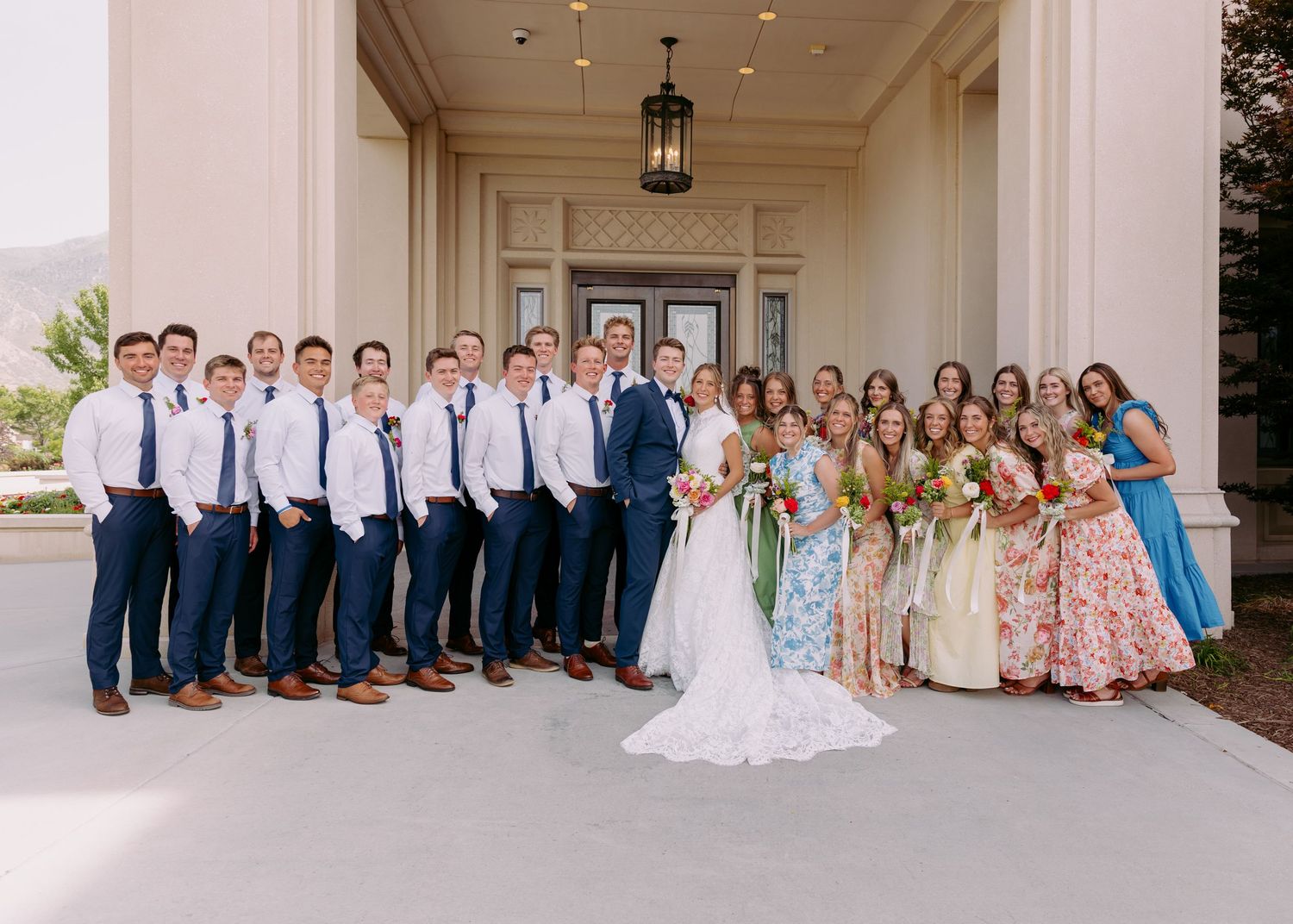 Wedding party poses outside venue with groomsmen in navy suits and bridesmaids in floral dresses flanking the bride and groom.