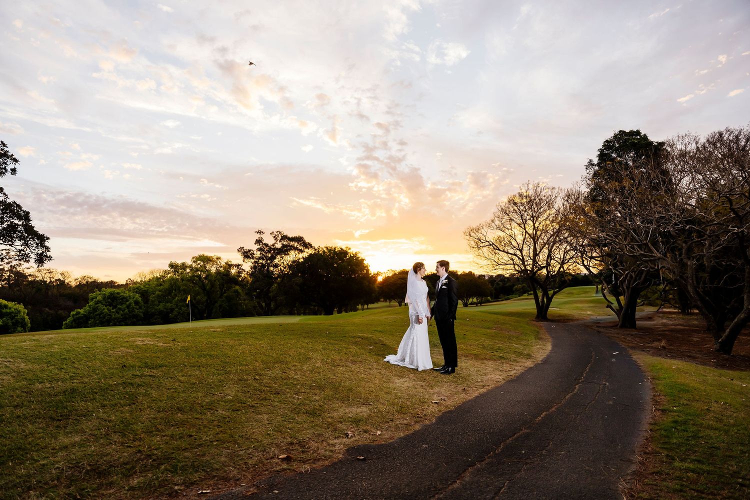 Bride and groom share a romantic moment on a winding path at sunset, surrounded by lush green trees at Indooroopilly Golf Club Queensland