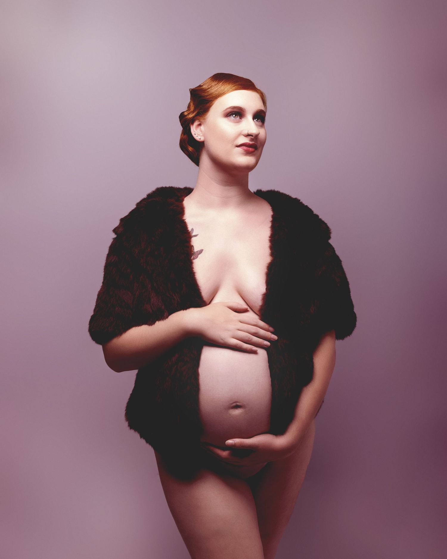 Pregnant redhead woman posing elegantly in fur wrap, cradling baby bump in artistic maternity portrait.