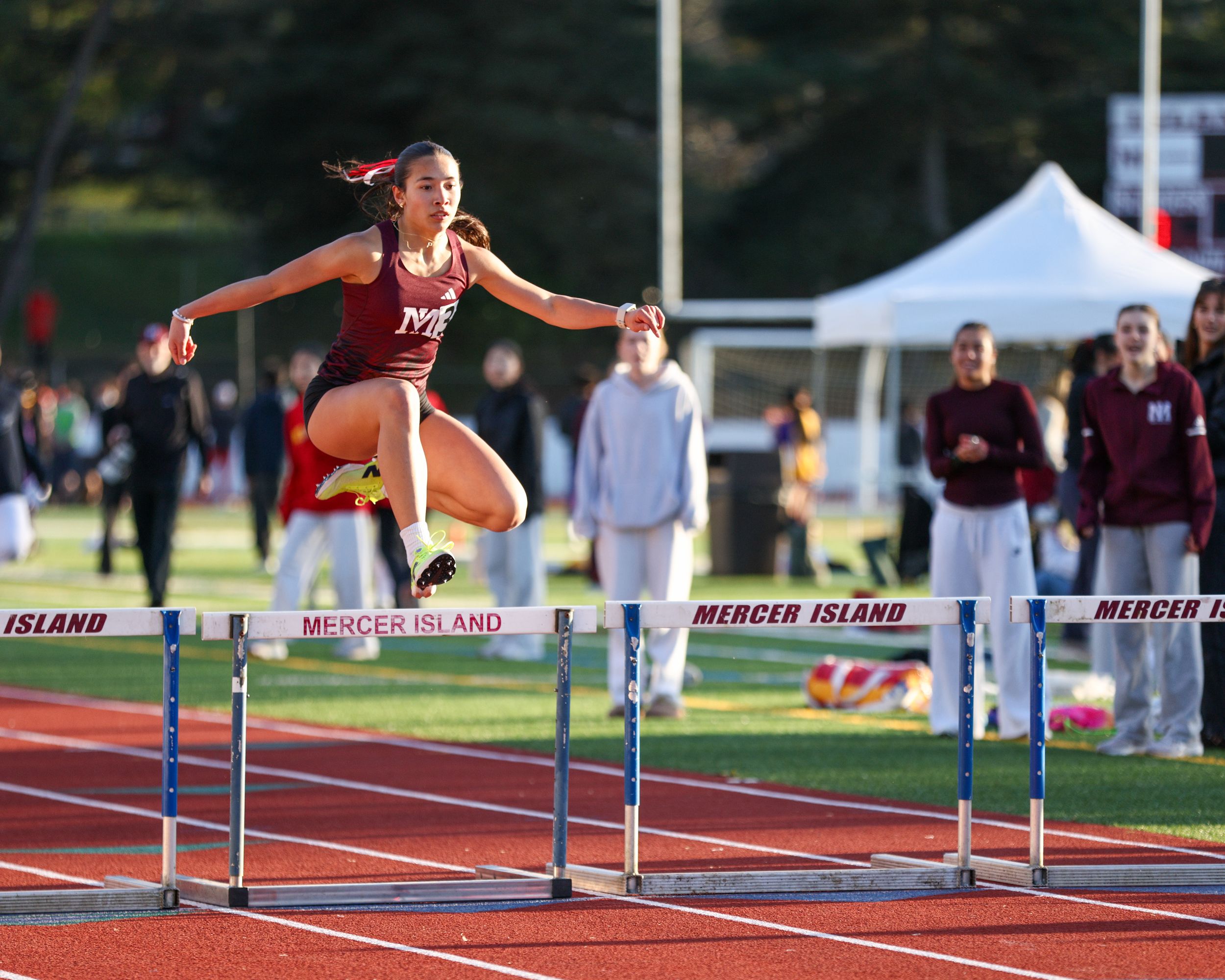 Female athlete clears a hurdle during a track and field competition at Mercer Island, mid-air in maroon uniform.