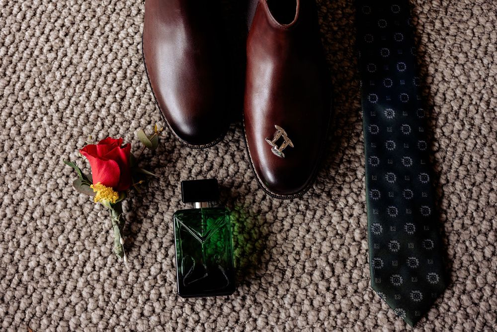 Brown leather dress shoes, boutonniere, green cologne bottle, and navy dotted tie arranged on gray carpet for a groom.