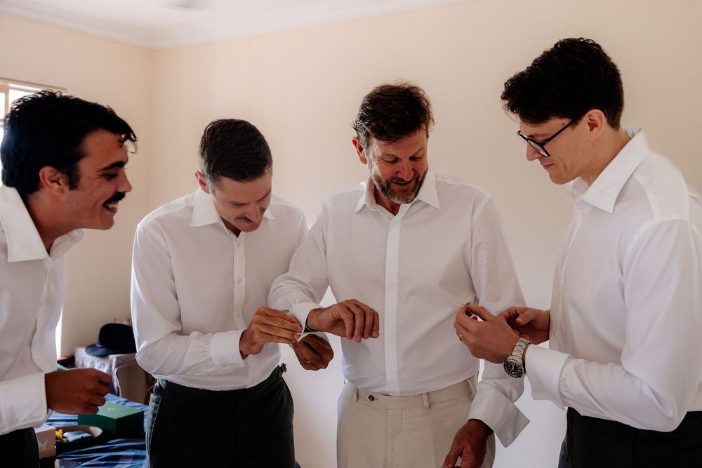 Four groomsmen in white shirts laughing and adjusting cufflinks while getting ready for a wedding.