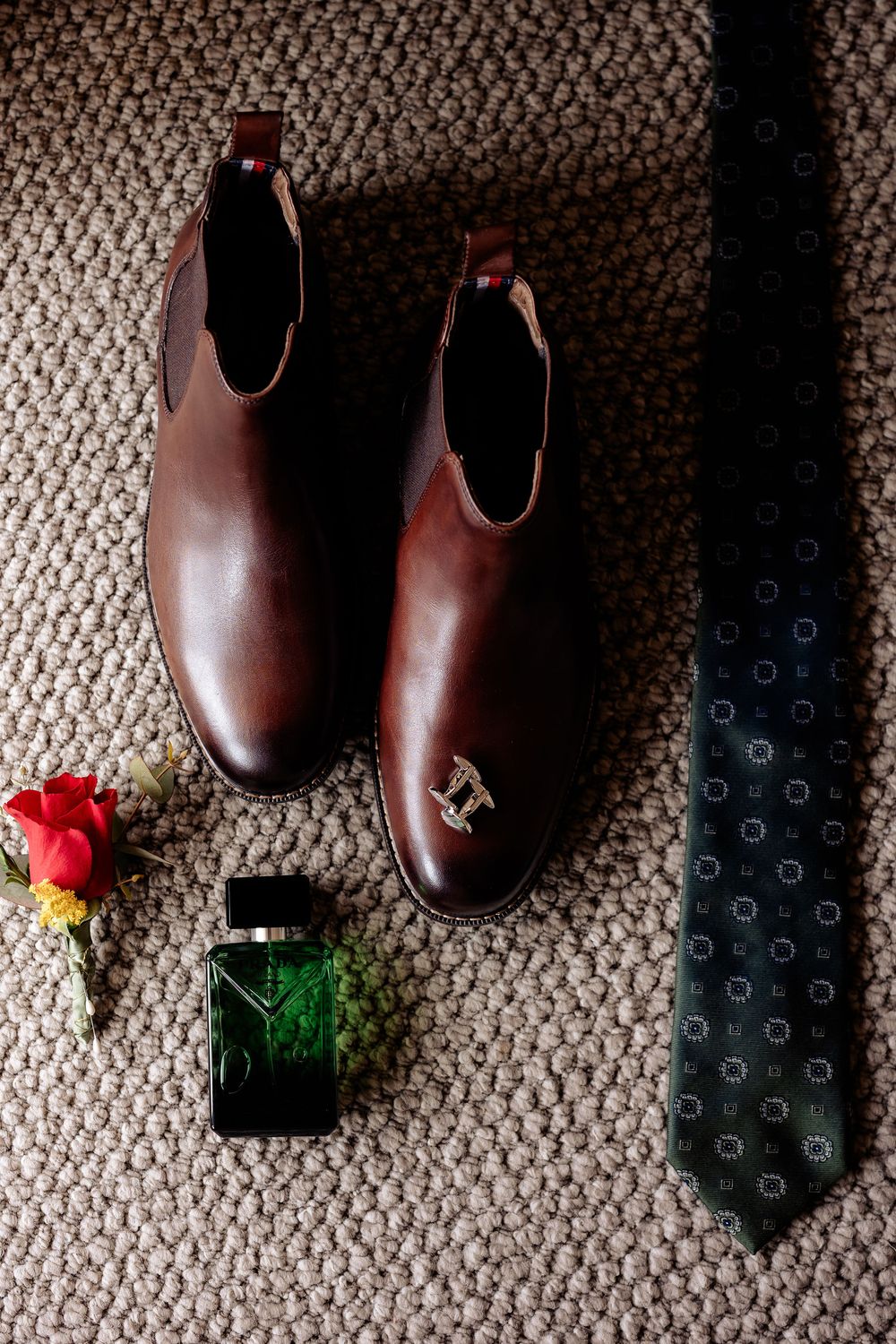 Brown leather Chelsea boots with a dark floral tie, green cologne bottle, and red rose boutonniere on a textured carpet.