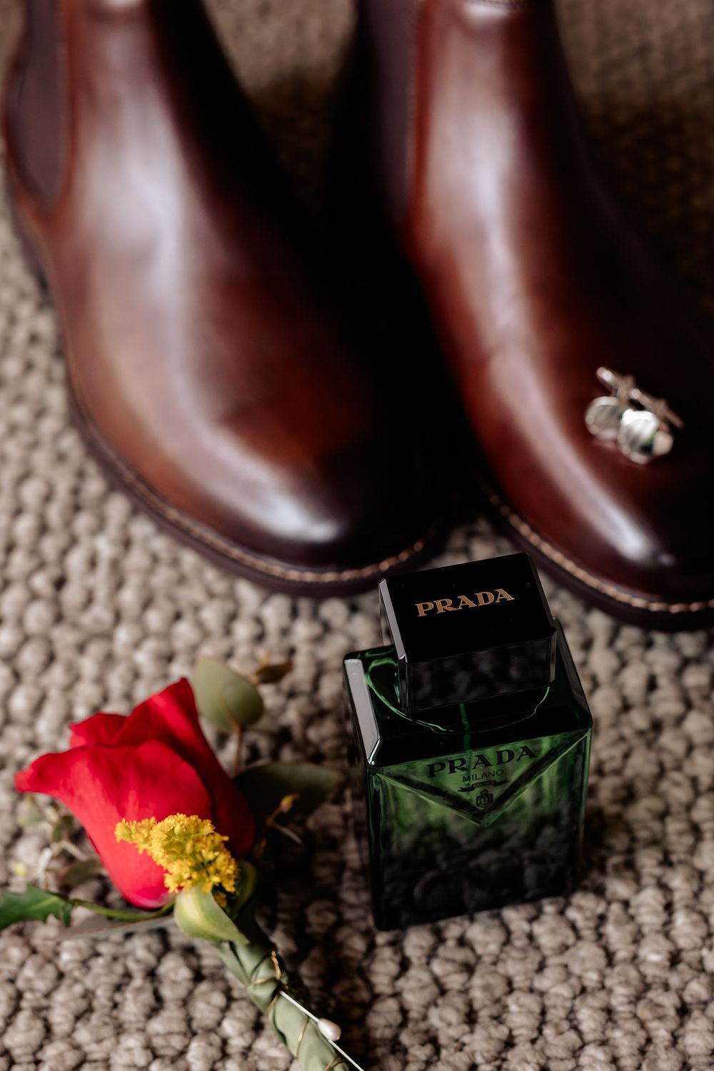Prada cologne bottle with red rose beside brown leather dress shoes and a bee lapel pin on textured carpet.
