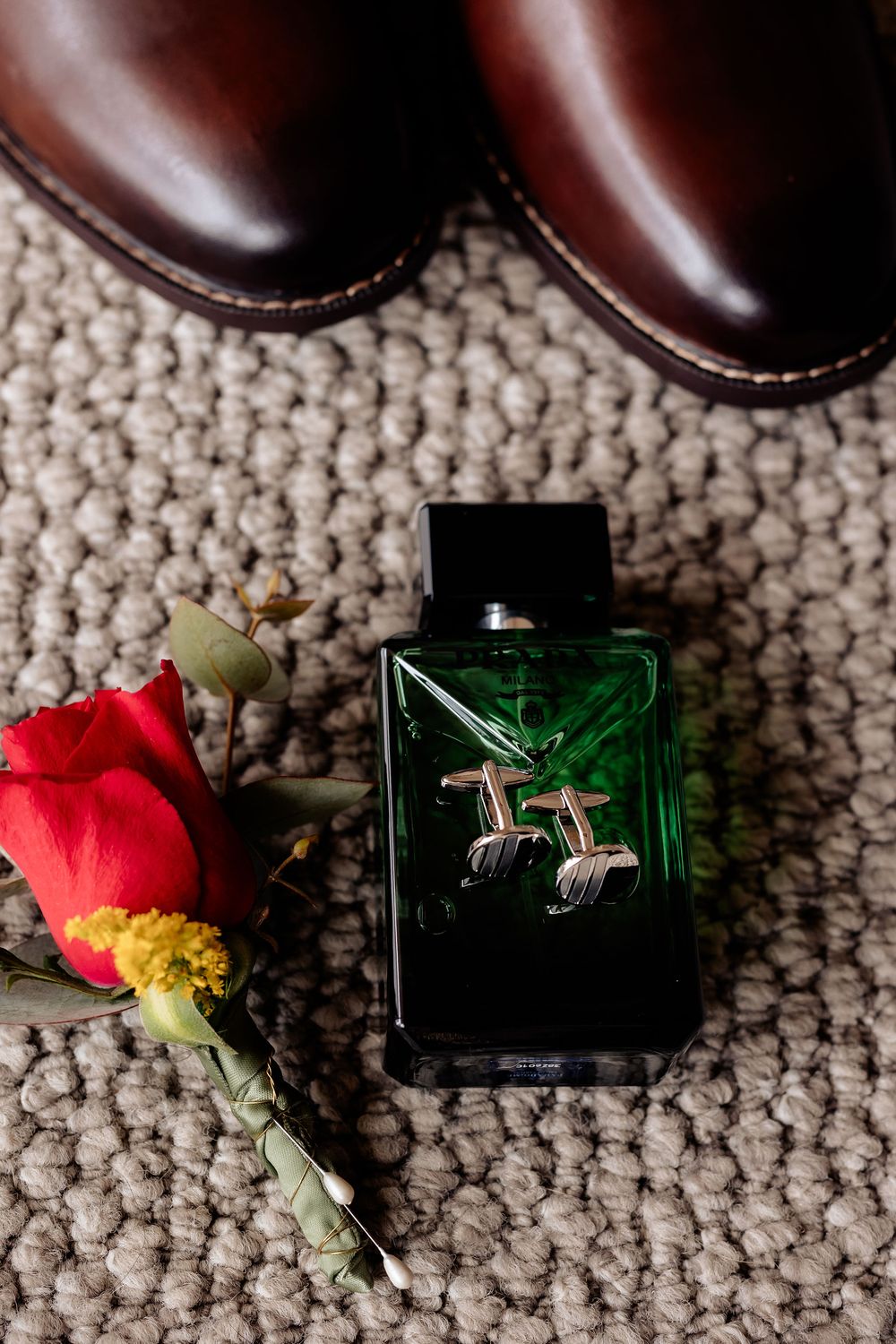 Close-up of silver cufflinks resting on a green cologne bottle beside a red rose boutonniere and brown leather dress shoes.