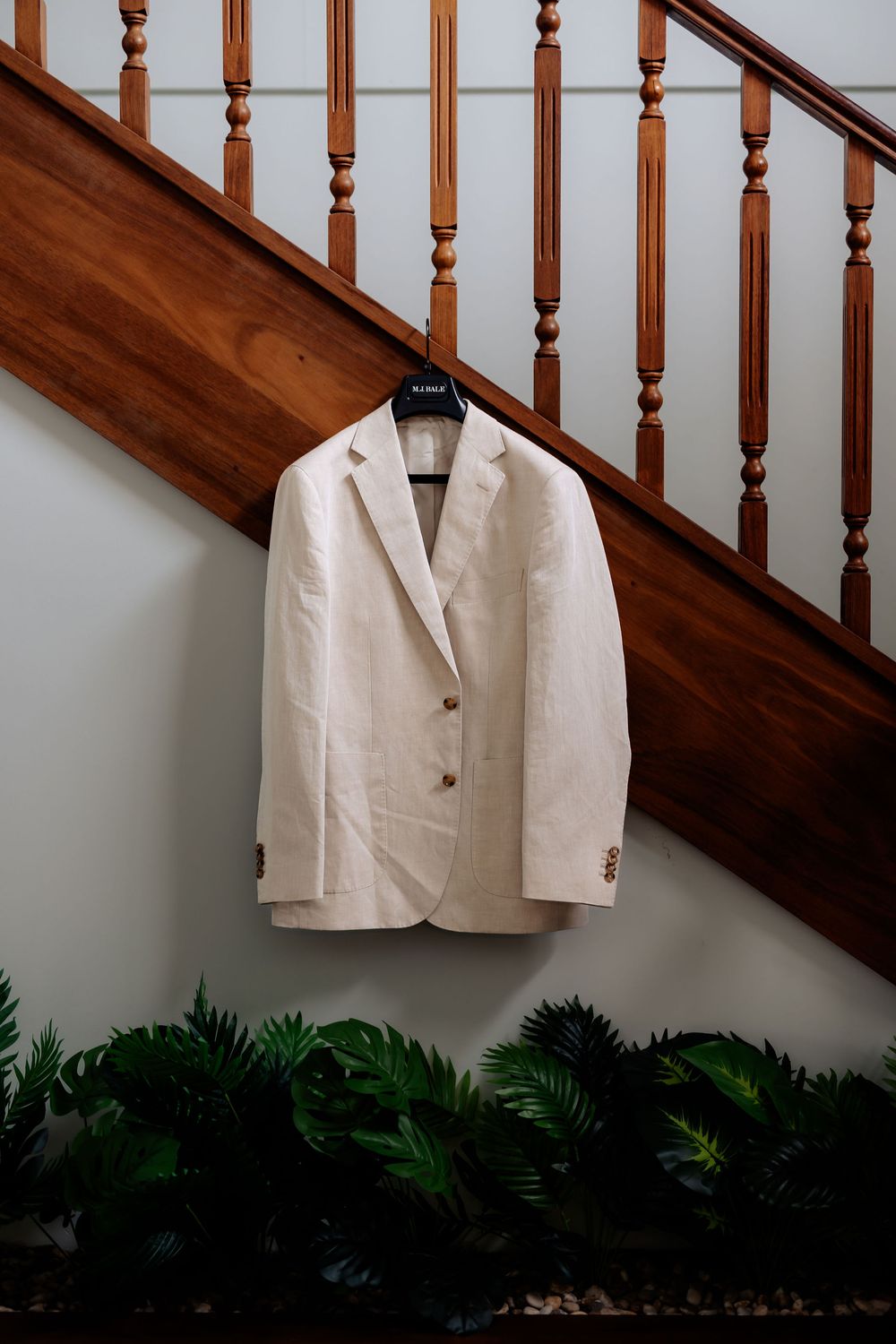 Cream linen blazer hanging on black hanger against staircase with wooden railing and tropical greenery below.