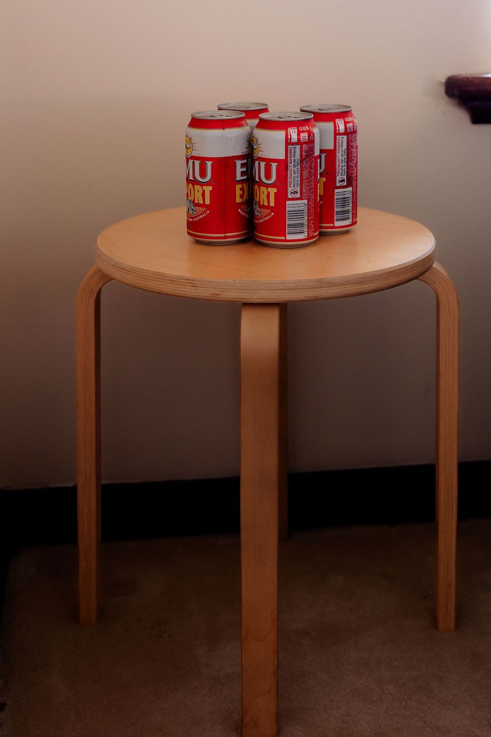 Four red Export beer cans sitting on a small round wooden three-legged stool against a beige wall.