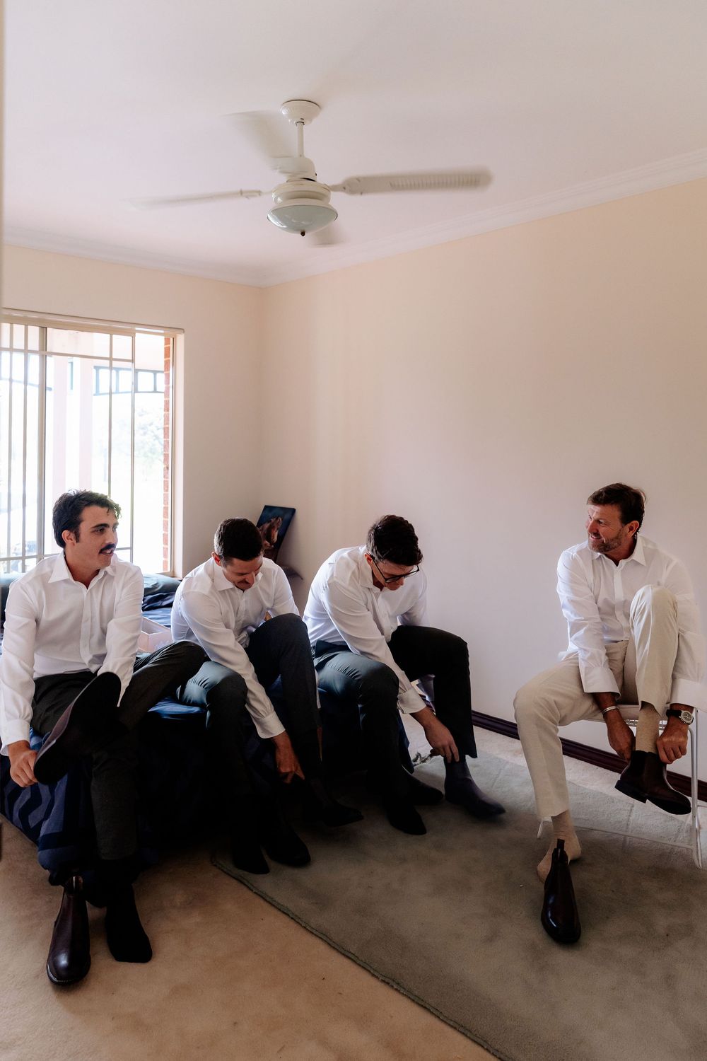 Four groomsmen in white shirts putting on black dress shoes while seated in a bright room with a ceiling fan.