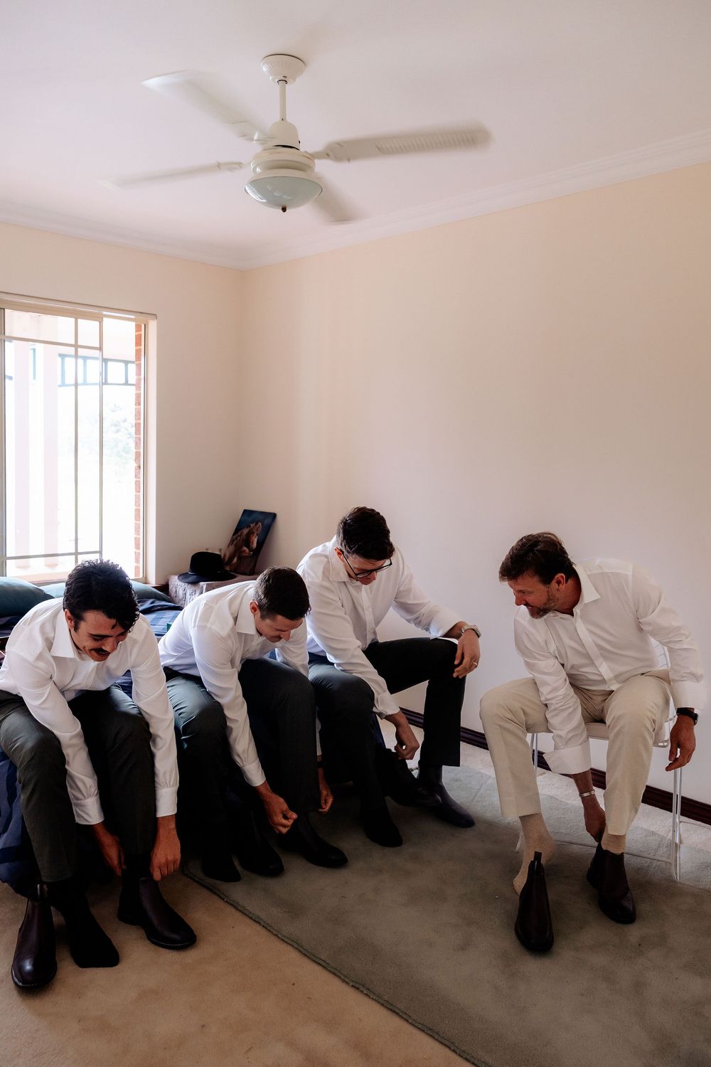 Four groomsmen in white shirts sitting together putting on dress shoes while getting ready for a wedding.
