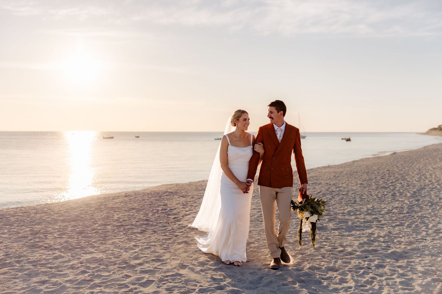 Bride and groom walking hand in hand on a sandy beach at sunset, groom in rust suit holding bridal bouquet.