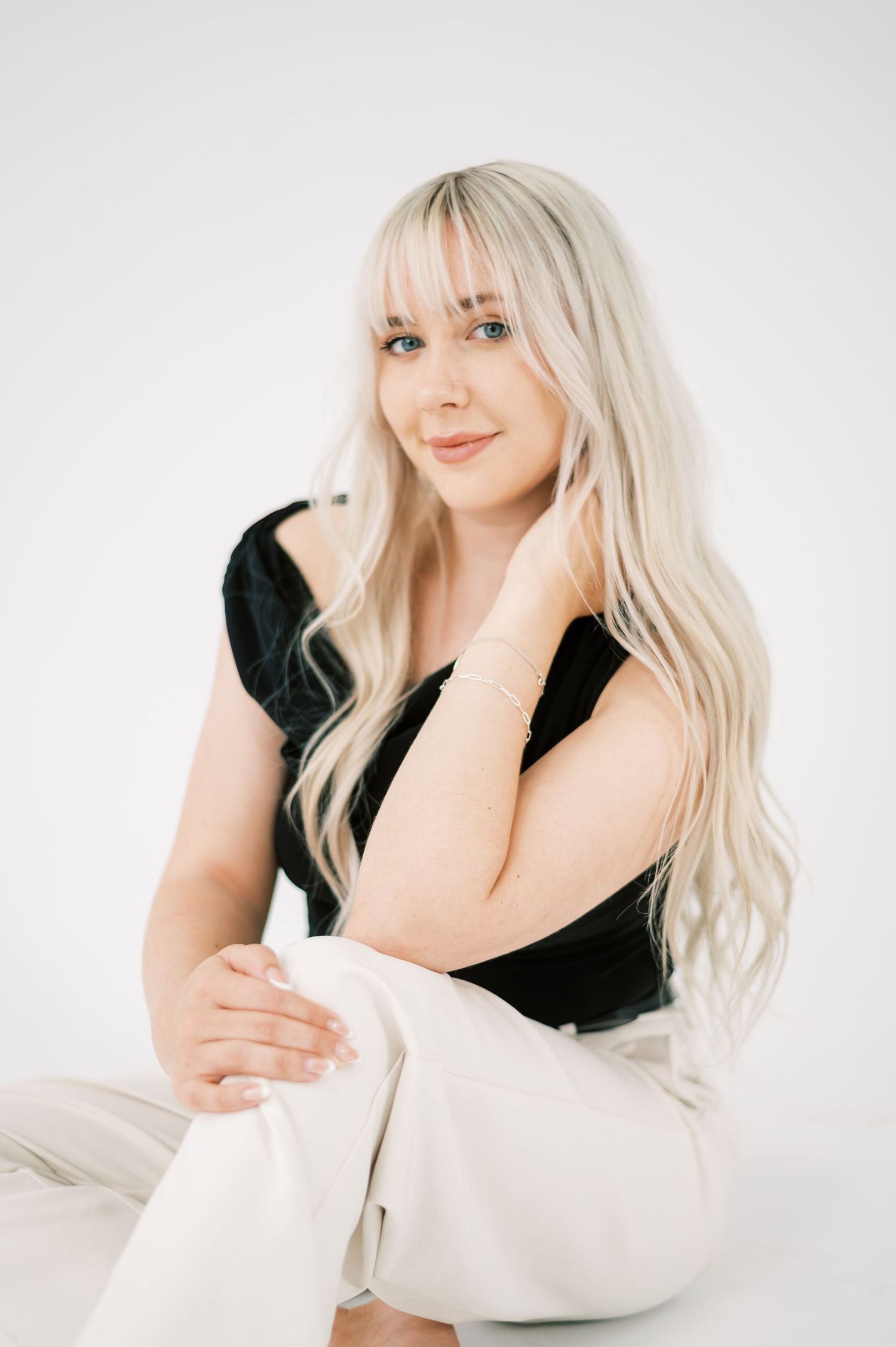 Young woman with long blonde hair sits gracefully in black top and white pants against white background.
