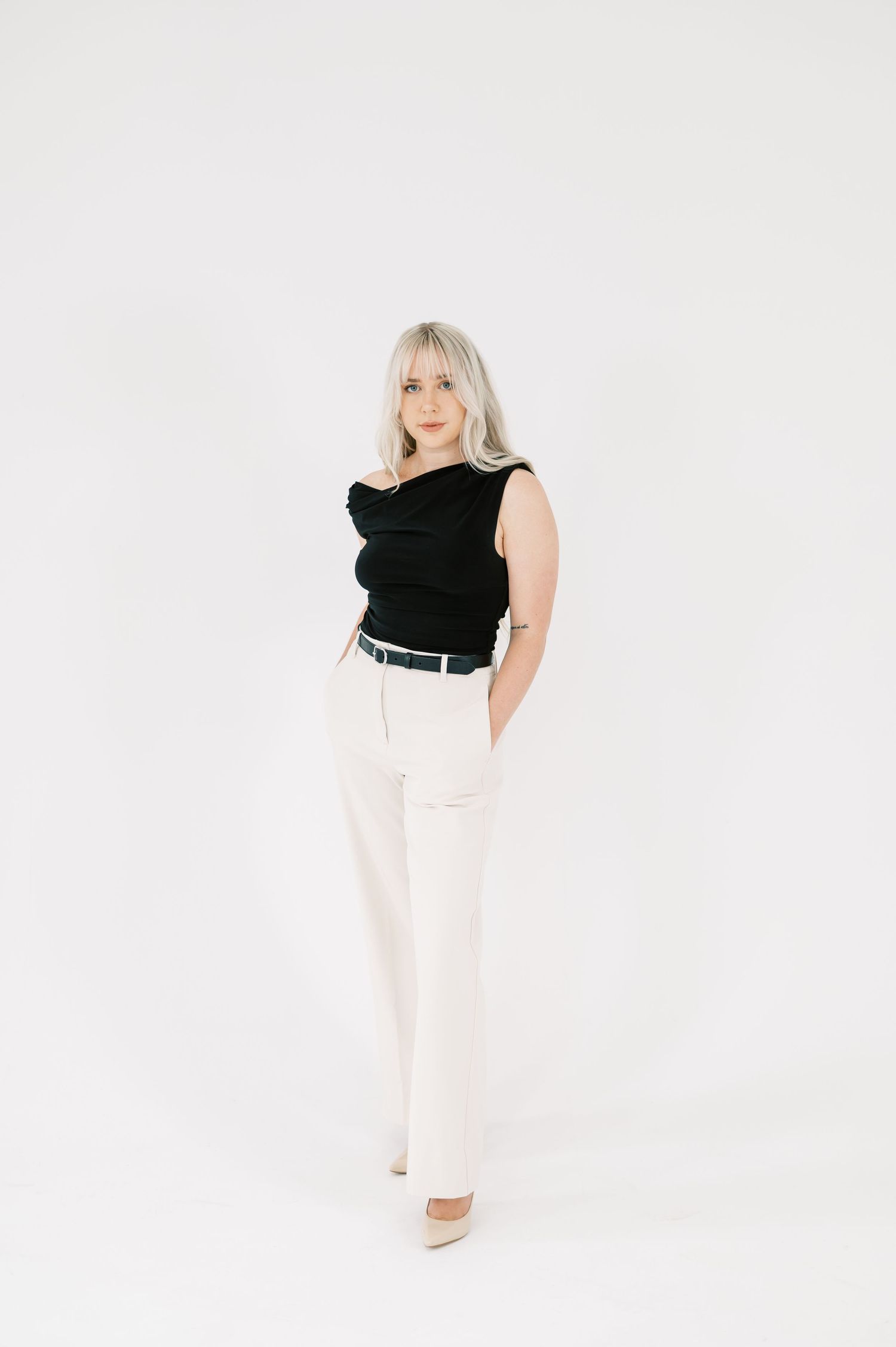 Blonde woman in black sleeveless top and white wide-leg trousers with belt, posing against white background.