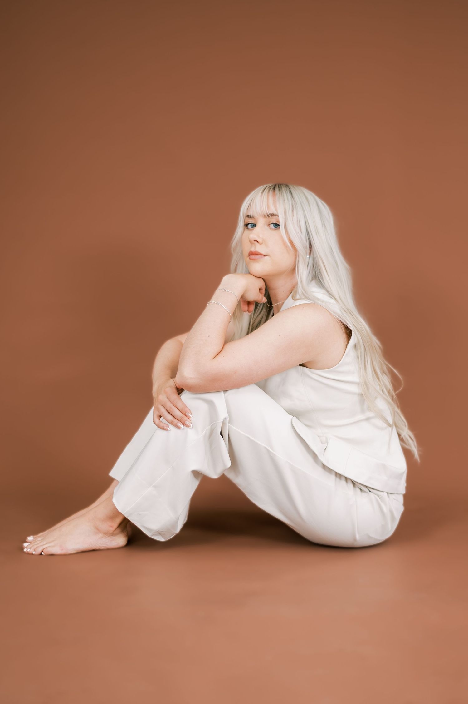 Young woman with long blonde hair wearing white outfit sitting barefoot on floor against brown background.