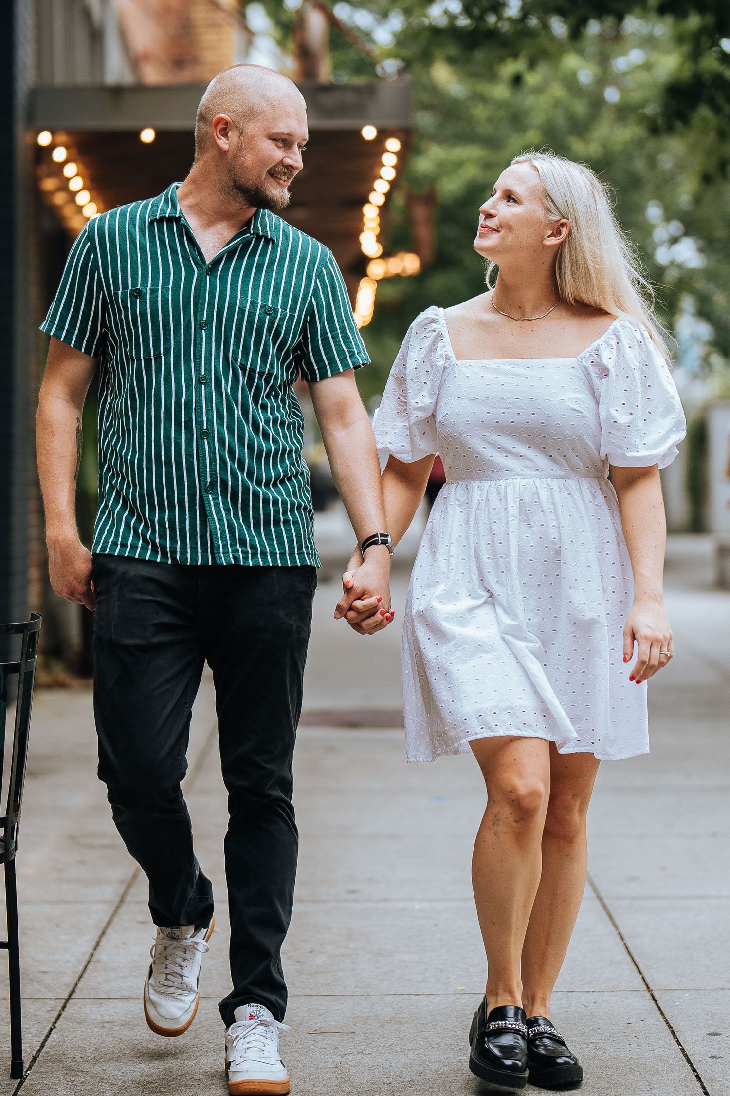 A couple walking hand in hand on a sidewalk, wearing a green striped shirt and white summer dress with string lights behind them.