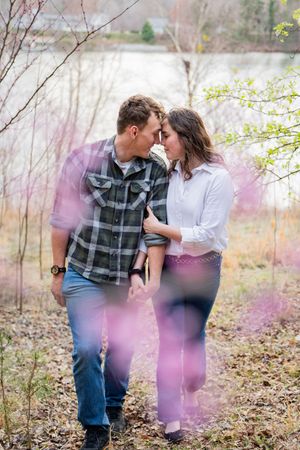 Couple sharing a romantic moment outdoors near a lake, surrounded by bare trees and soft pink bokeh light.