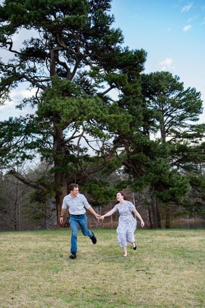A joyful couple runs hand-in-hand across a green lawn with tall pine trees in the background.
