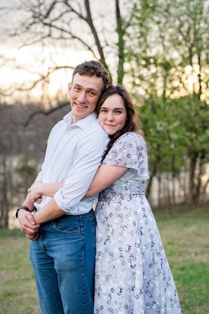 A young couple embraces outdoors at sunset, woman in floral dress hugging man in white shirt and jeans, trees in background.