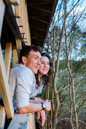 Smiling young couple leaning on wooden balcony railing surrounded by bare trees outdoors.