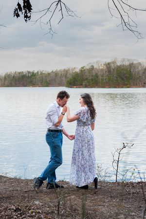 Romantic couple dancing by a serene lake, man kissing woman's hand, wearing floral dress, trees reflecting on calm water.