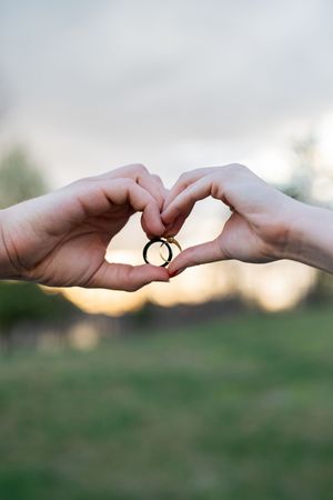 Two hands forming a heart shape holding wedding rings outdoors at sunset.