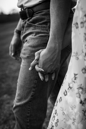 Black and white photo of a couple holding hands, woman wearing floral dress with manicured nails, man in jeans with belt.