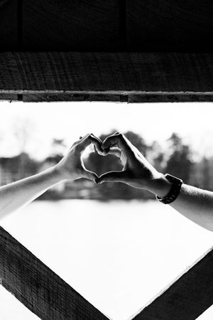 Black and white photo of two people forming a heart shape with their hands together near a wooden window frame.