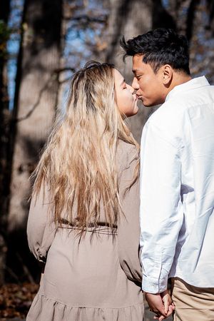A romantic couple in casual attire shares an intimate moment while standing together in a wooded outdoor setting.