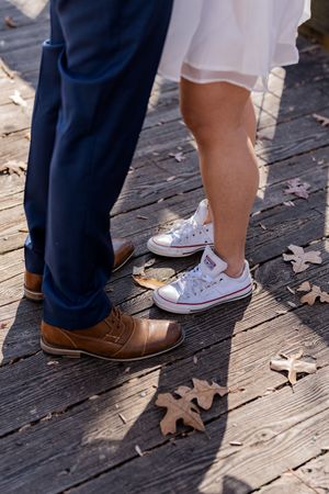 Close-up of feet showing brown leather dress shoes and white sneakers on a wooden deck with scattered autumn leaves.