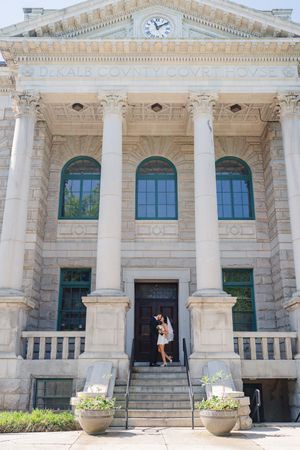 Couple kissing on steps of DeKalb County Courthouse, a grand neoclassical building with tall columns and arched windows.