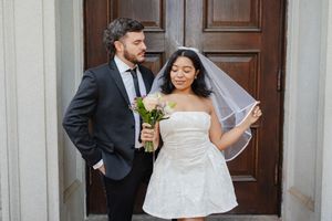 Bride in white mini dress and veil holds pink bouquet beside groom in black suit at wooden door.