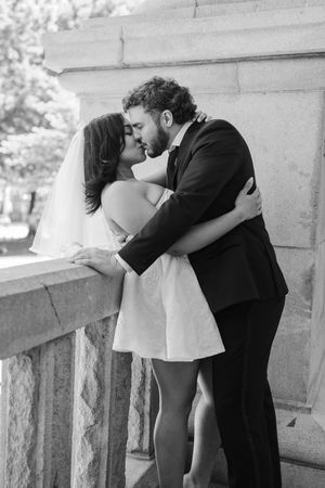 Black and white photo of a bride and groom sharing a romantic kiss against a stone pillar on their wedding day.