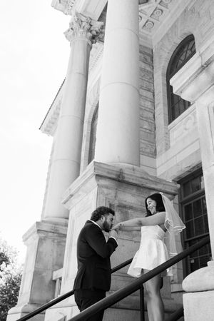 Black and white photo of groom kissing bride's hand on steps of elegant building with tall columns.