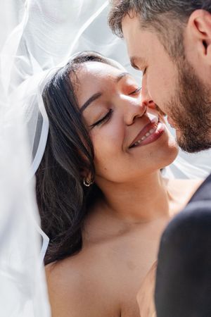 A smiling bride with closed eyes leans close to her bearded groom under a white wedding veil.