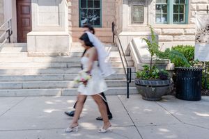 Bride in short white dress and veil walks with companion outside stone building on wedding day.