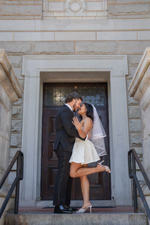 Bride and groom share romantic kiss on church steps, bride in mini dress and veil lifting one leg.