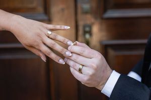 Close-up of a couple exchanging gold wedding rings, bride with manicured nails, groom in suit, wooden door background.