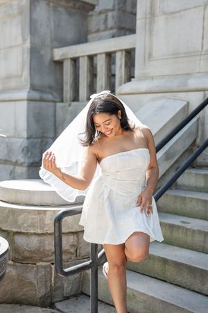Smiling bride in white strapless mini dress and veil poses on stone steps outdoors.