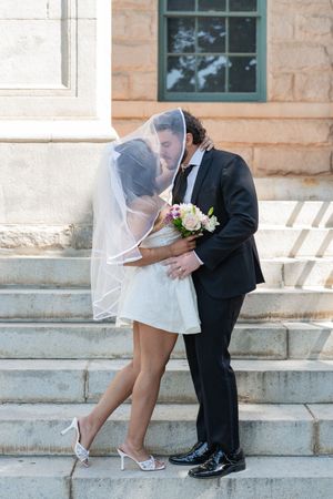 Newlyweds share a kiss on stone steps, bride in short white dress and veil holding a bouquet.