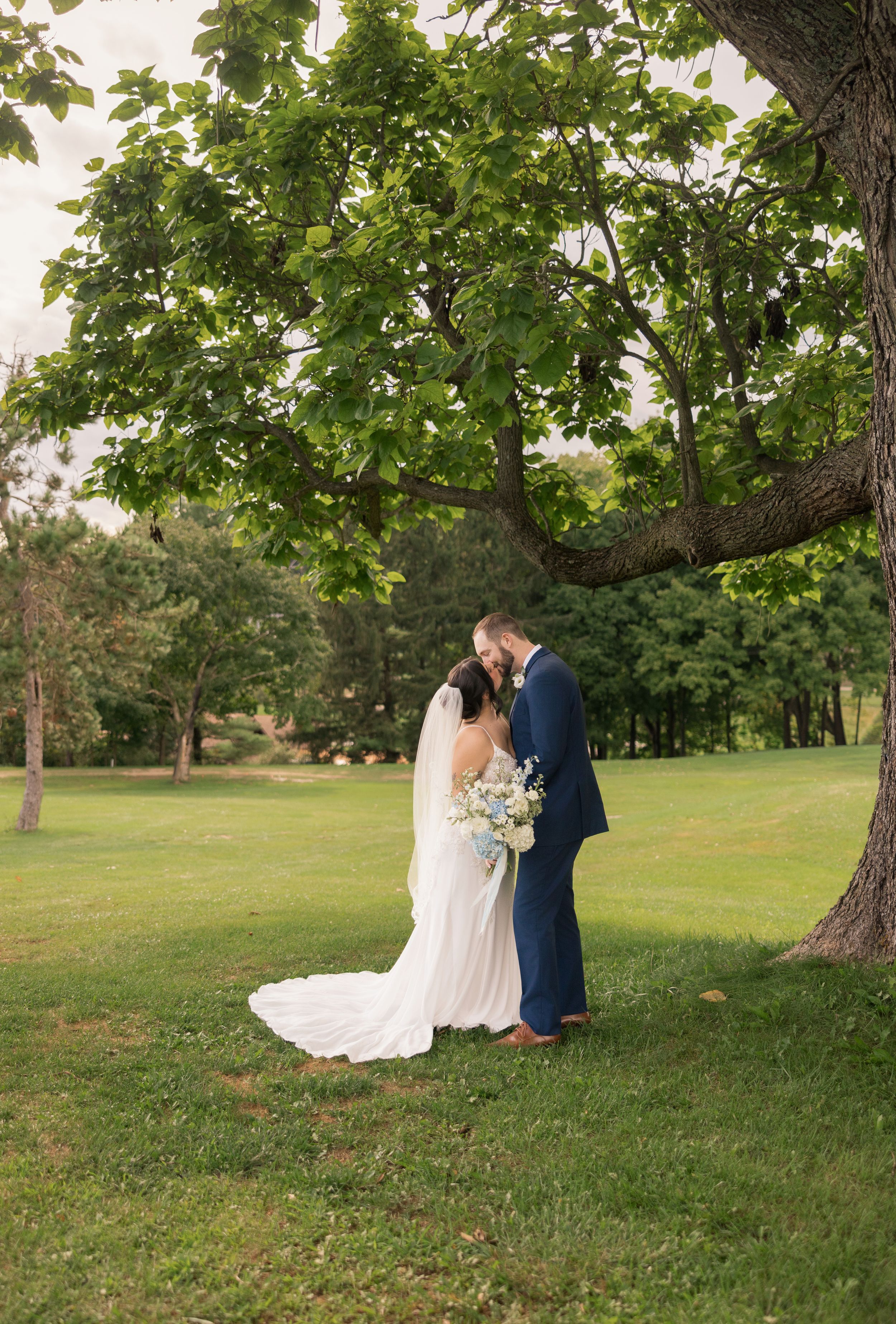 Bride and groom share a romantic moment under a large tree on a lush green lawn during their outdoor wedding.