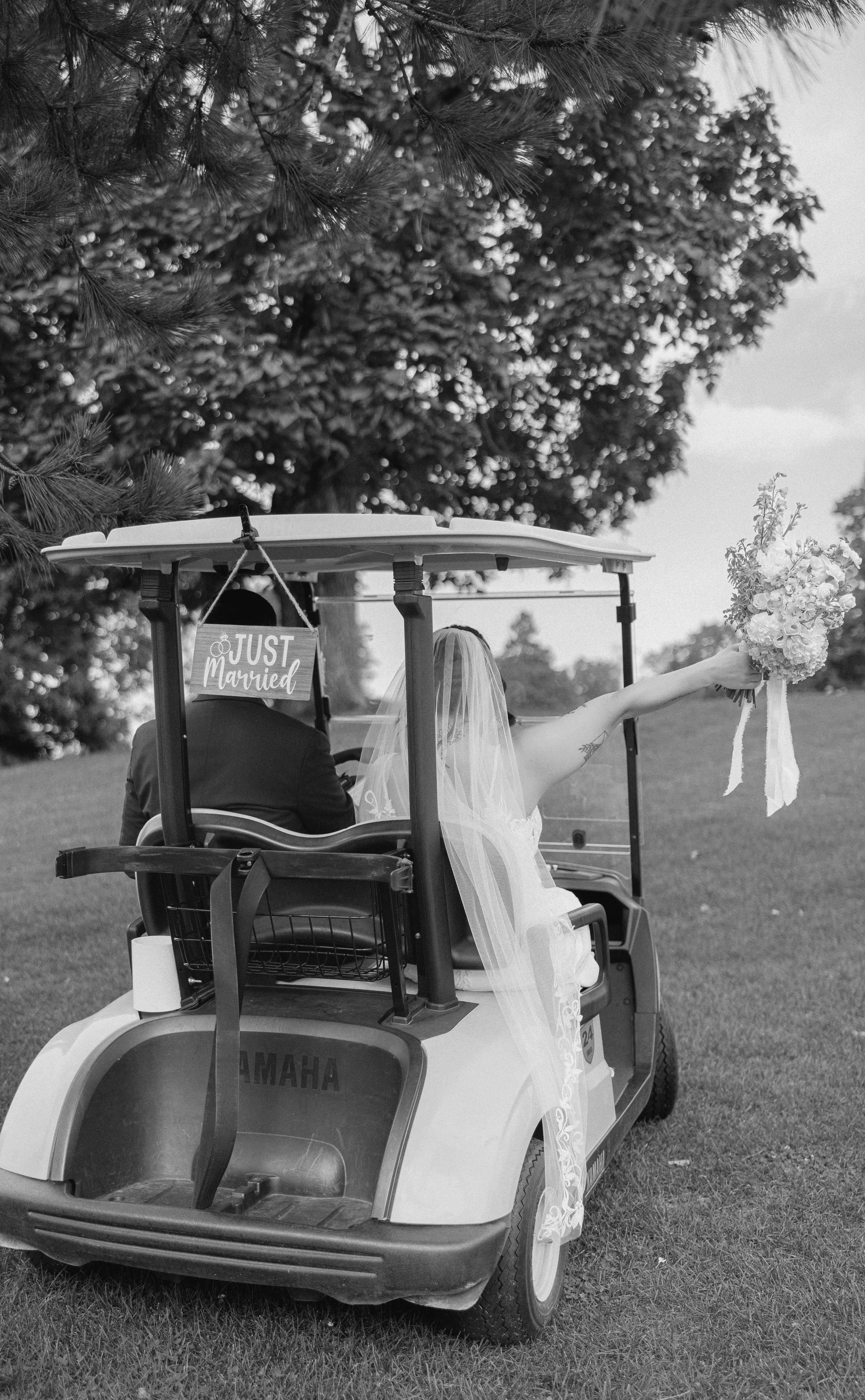 Newlyweds ride a Yamaha golf cart with a Just Married sign, bride holding bouquet aloft, in black and white wedding photo.
