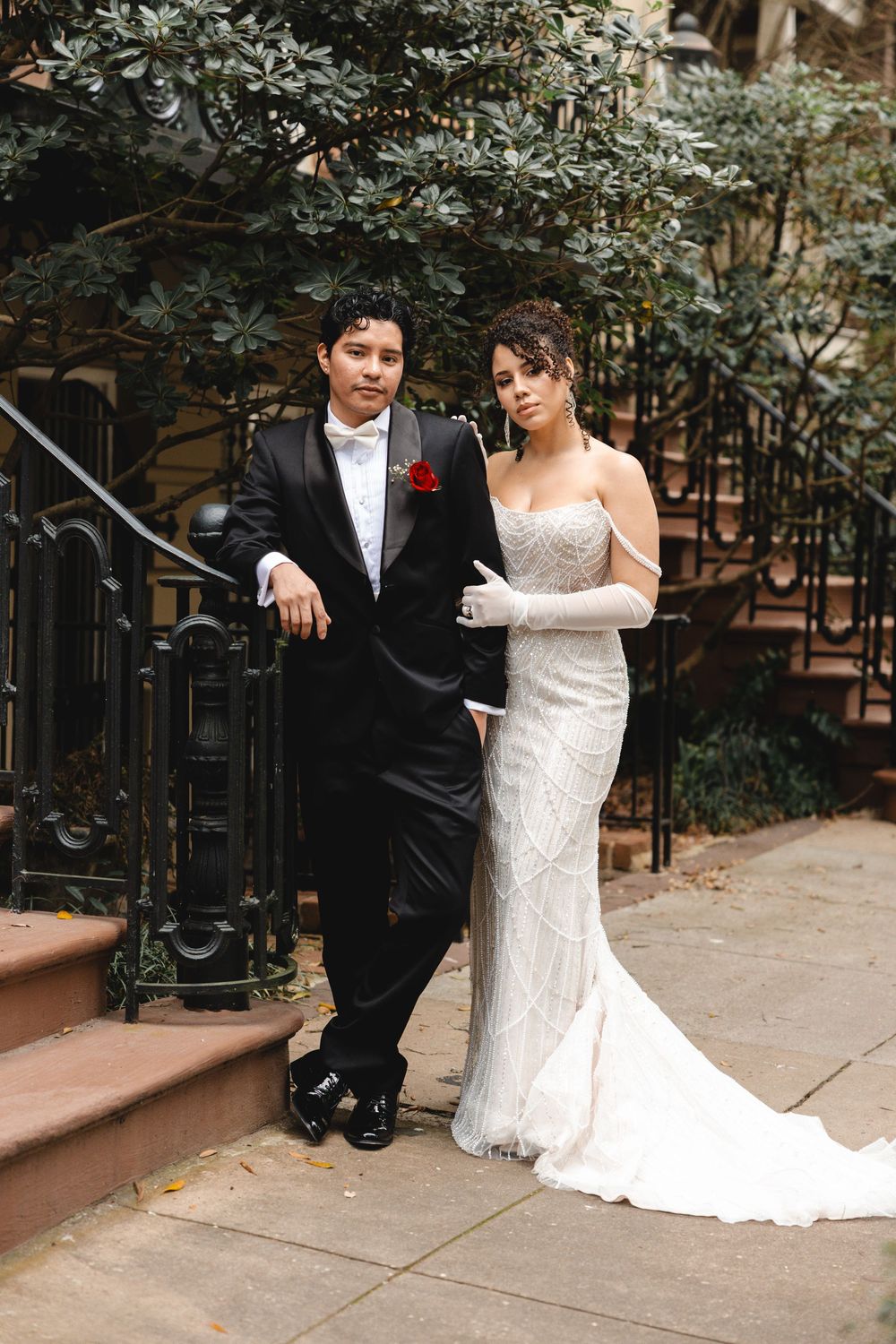 Bride in beaded gown with gloves and groom in black tuxedo posing outdoors on urban steps with lush greenery.